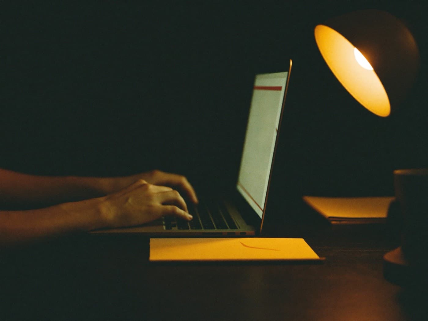 Mãos digitando em um notebook sob a luz de um abajur